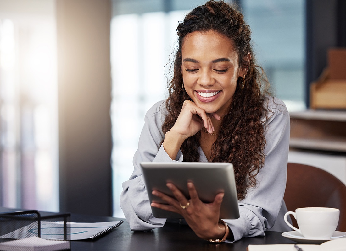 Read Our Reviews - Happy Young Woman Reading a Tablet While Sitting at Her Kitchen Table With a Cup of Coffee